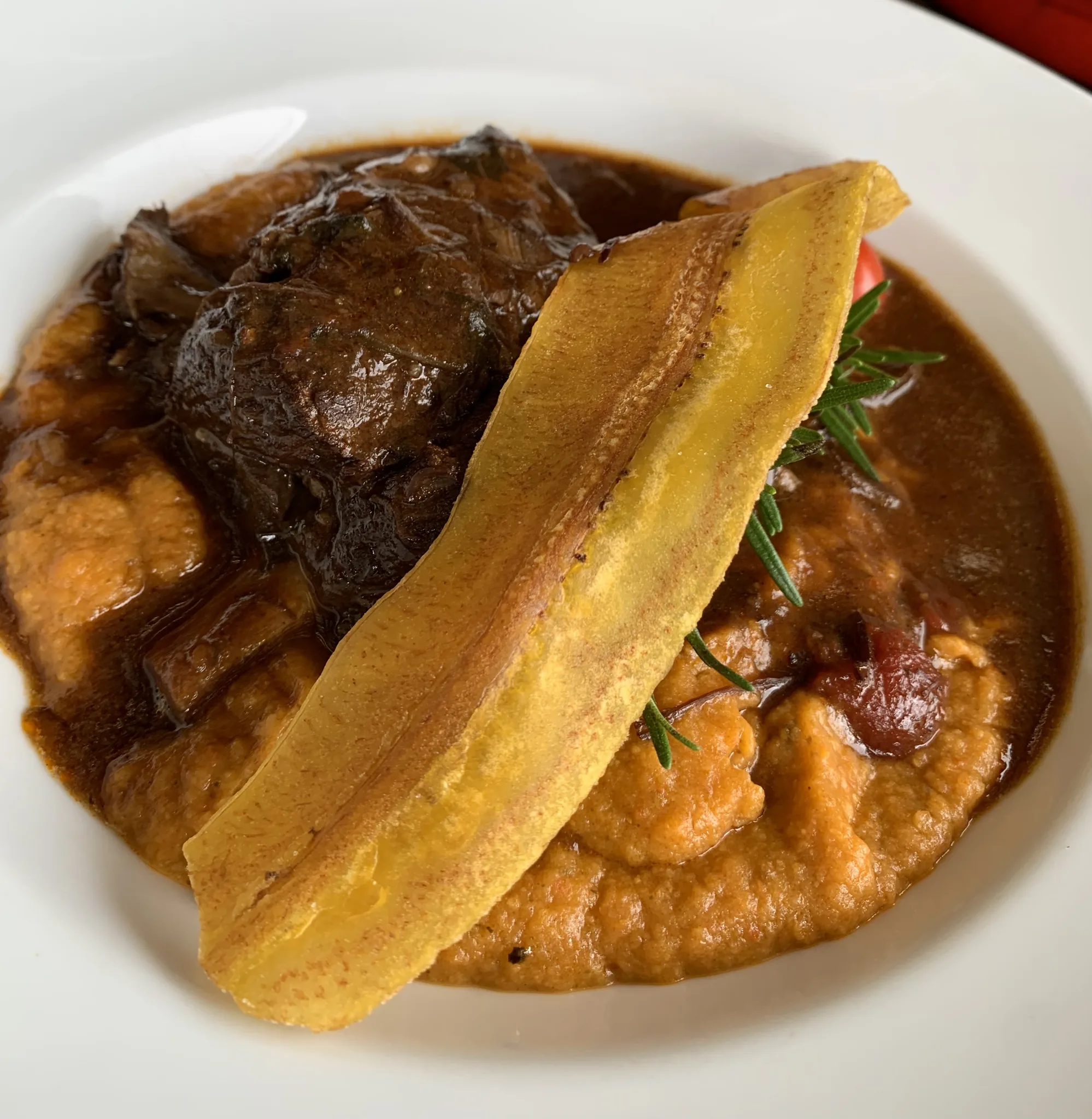 Costilla guisada sobre un espiral de puré de camote, con una tajada de plátano frito y una ramita de romero.