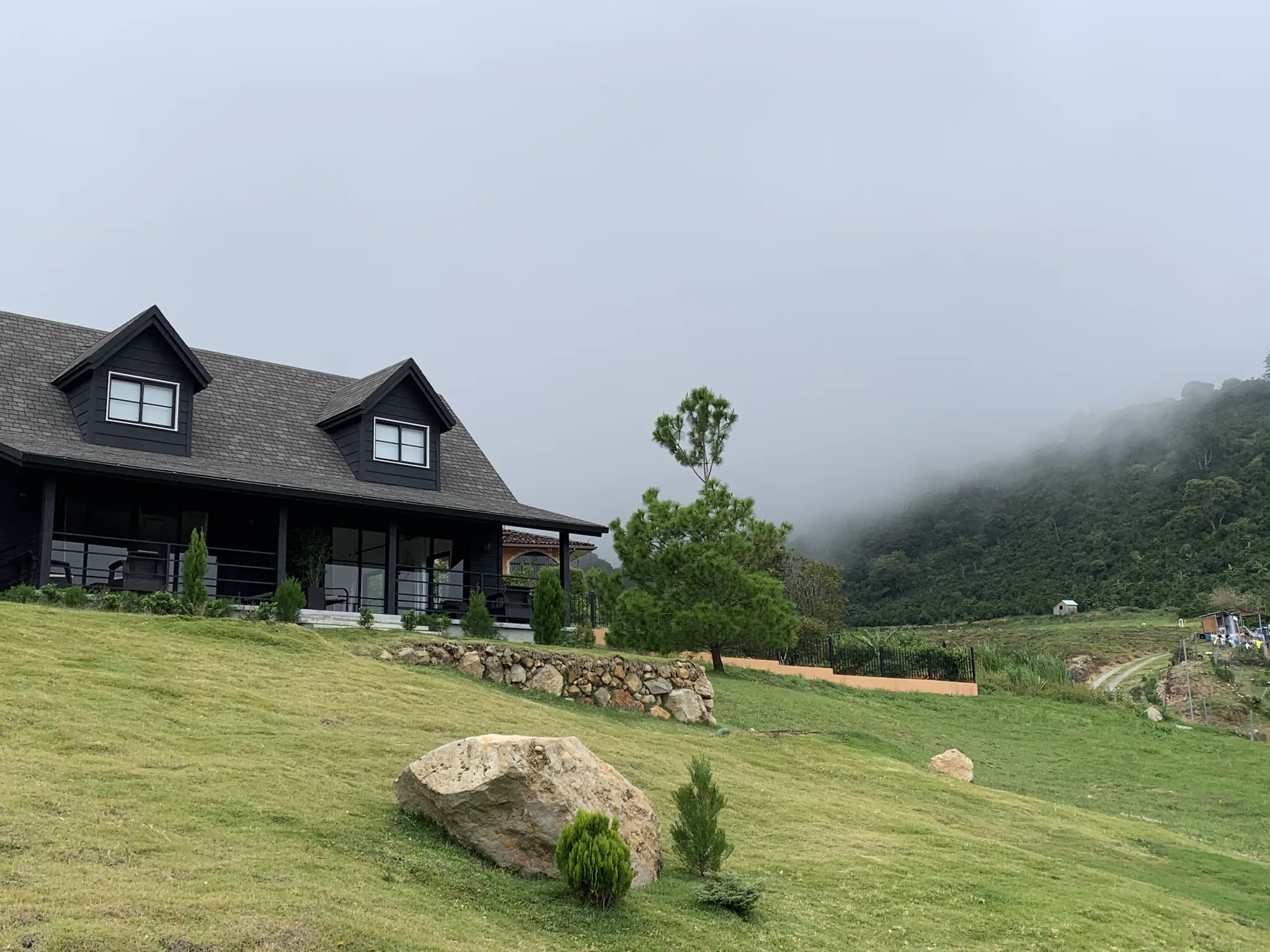 A dark cabin with dormer windows and a stone retaining wall sits on a mowed lawn as fog rolls over the forested ridge behind it.