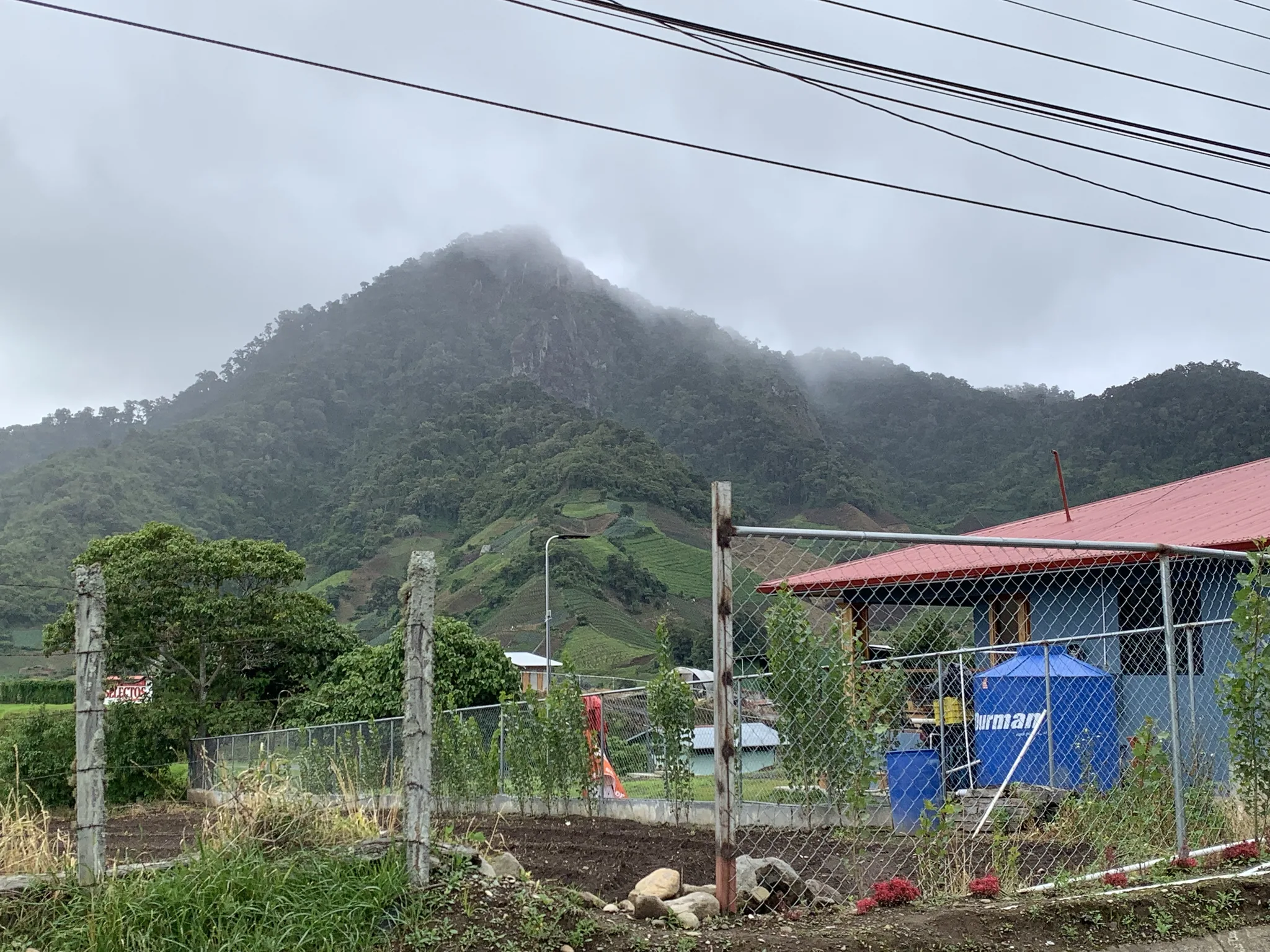 Un cerro envuelto en nubes se levanta detrás de una cerca de malla, parcelas en terraza y una casa azul de techo de zinc rojo.